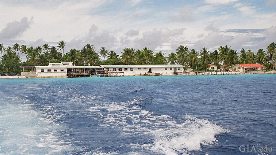 Landscape of French Polynesia's atolls shielding pearl farms, where the June birthstone is found, from surrounding ocean waves. The land is lined with palm trees, sheltering a lagoon that's ideal for mollusk culturing. 