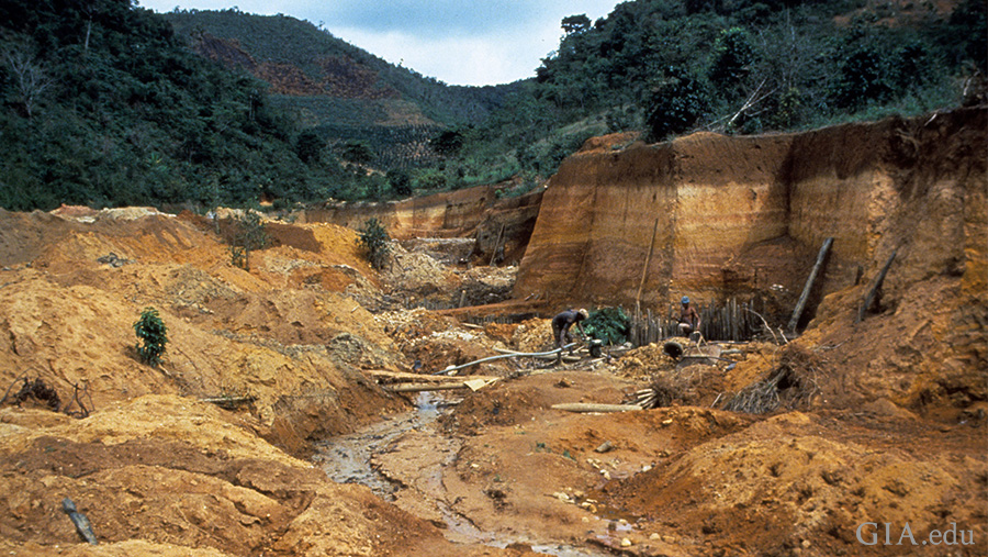 Landscape of a carved out mining ditch with a small stream running through, surrounded by tree-covered mountains in Brazil where the June birthstone, alexandrite, is found.