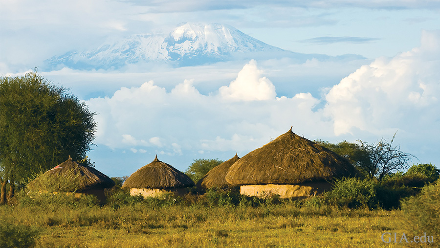 Landscape of huts in a field with the summit of Mount Kilimanjaro emerging from the clouds in the distance, where the December birthstone tanzanite is mined.